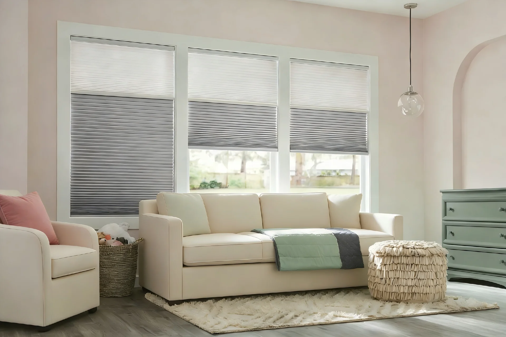Living room with pleated blinds, beige sofa, armchair, wicker basket, and pouf, accented by green dresser and pendant light.