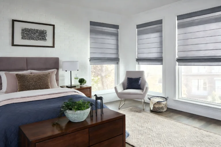 Modern bedroom featuring a bed with layered pillows, nightstand with lamp and plant, light gray chair with cushion, and large windows with gray shades.