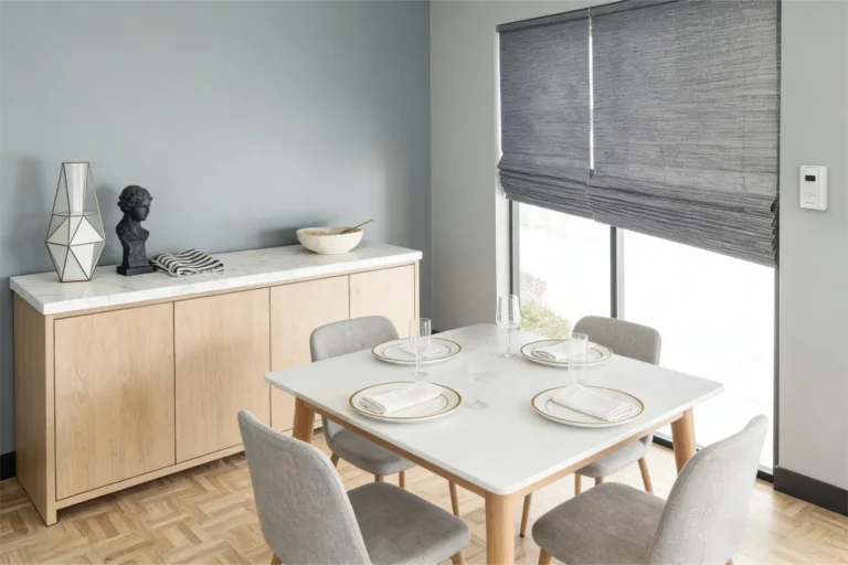 Modern dining room featuring a white table set with plates and glasses, surrounded by gray chairs. A sideboard is adorned with decor items and a bowl, with large windows covered by gray blinds.