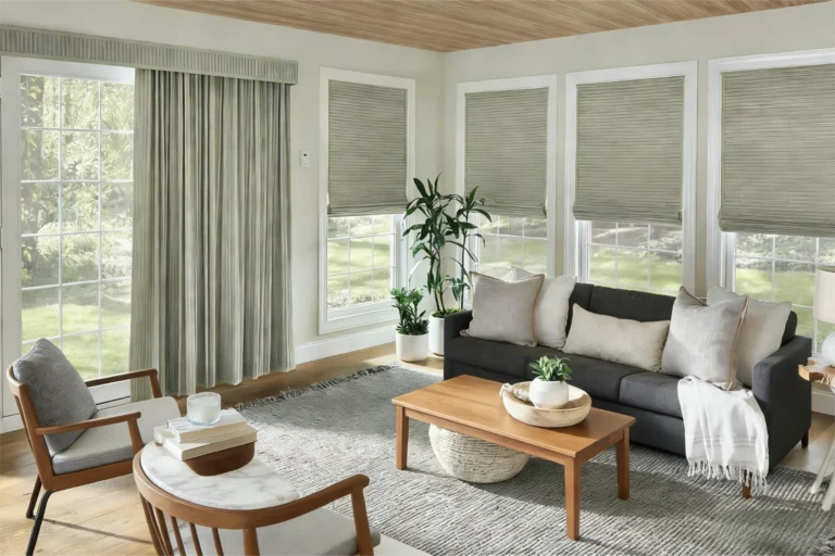 Living room featuring a dark gray sofa with pillows, wooden coffee table, armchairs, large windows with blinds, and green plants.