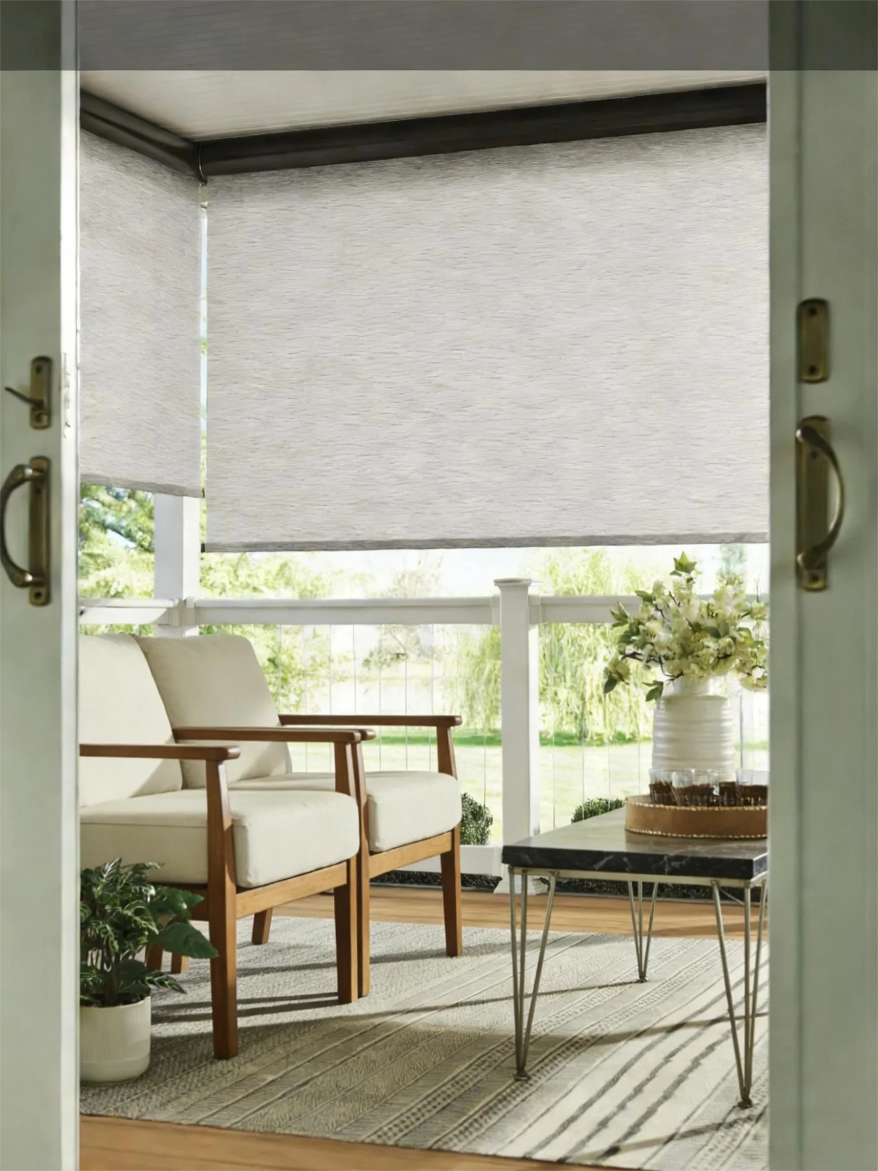 Open porch with beige chairs, black table, decorative plants, and roller blinds partially down, viewed from doorway.