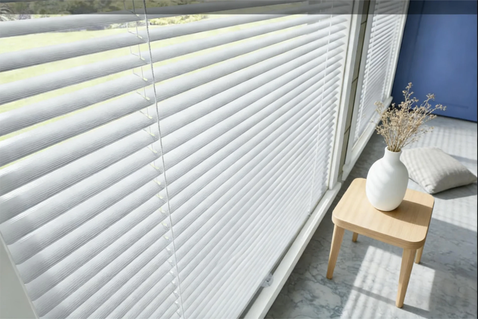 White blinds covering large windows beside a wooden table with a white vase and dried flowers. Light filters through with a cushion nearby.