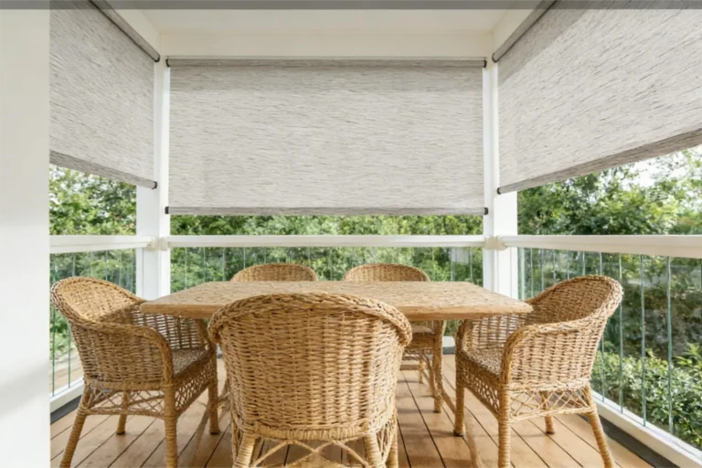 Woven wicker chairs and table set on a wooden deck with surrounding trees and pull-down blinds creating a cozy outdoor space.