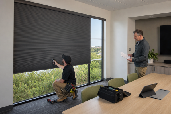 Technician commissioning a graphite motorized cellular shade while a project manager reviews the handover sheet