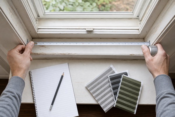 Hands measuring an older window opening beside gray and moss-green shade samples