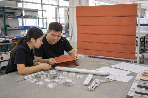 Factory staff reviewing a terracotta Roman shade sample, motor parts, cables, and a bill of materials